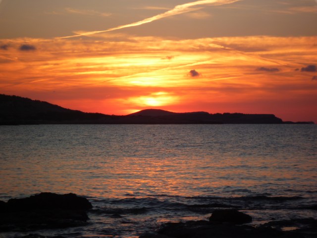 The red and orange sky of a sunset seen from across a bay.