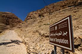A ridge of bare rock under a blue sky. In the foreground is a sign written in Arabic script and English saying "Mount of Temptation".d