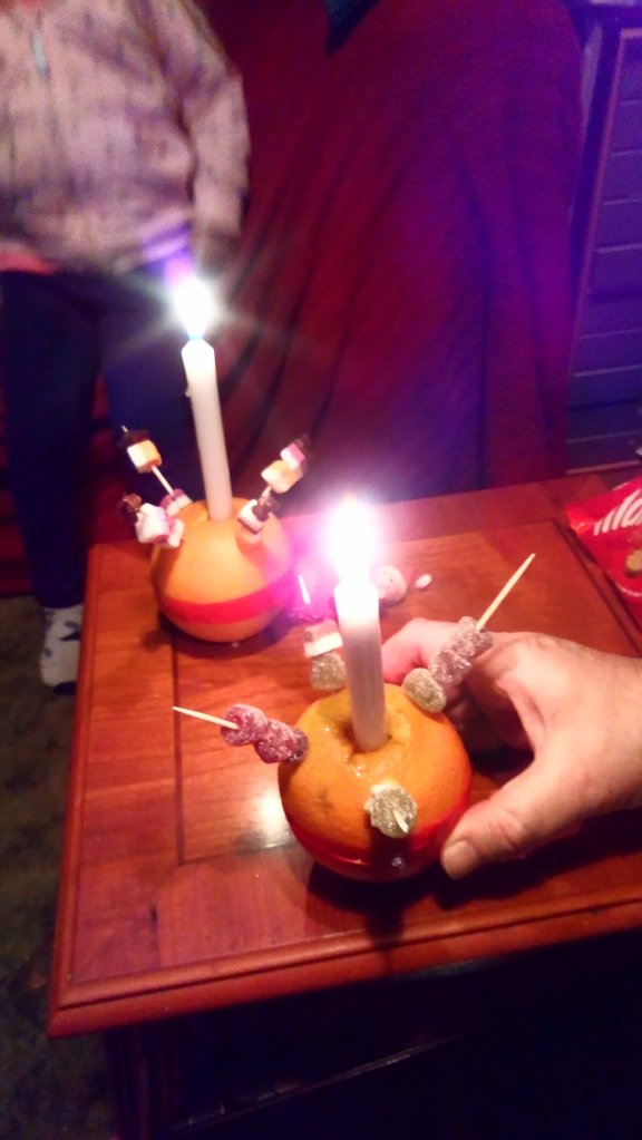 Christingles. Oranges with a lit candle in the centre, a red band around the middle and sweets on coctail sticks.