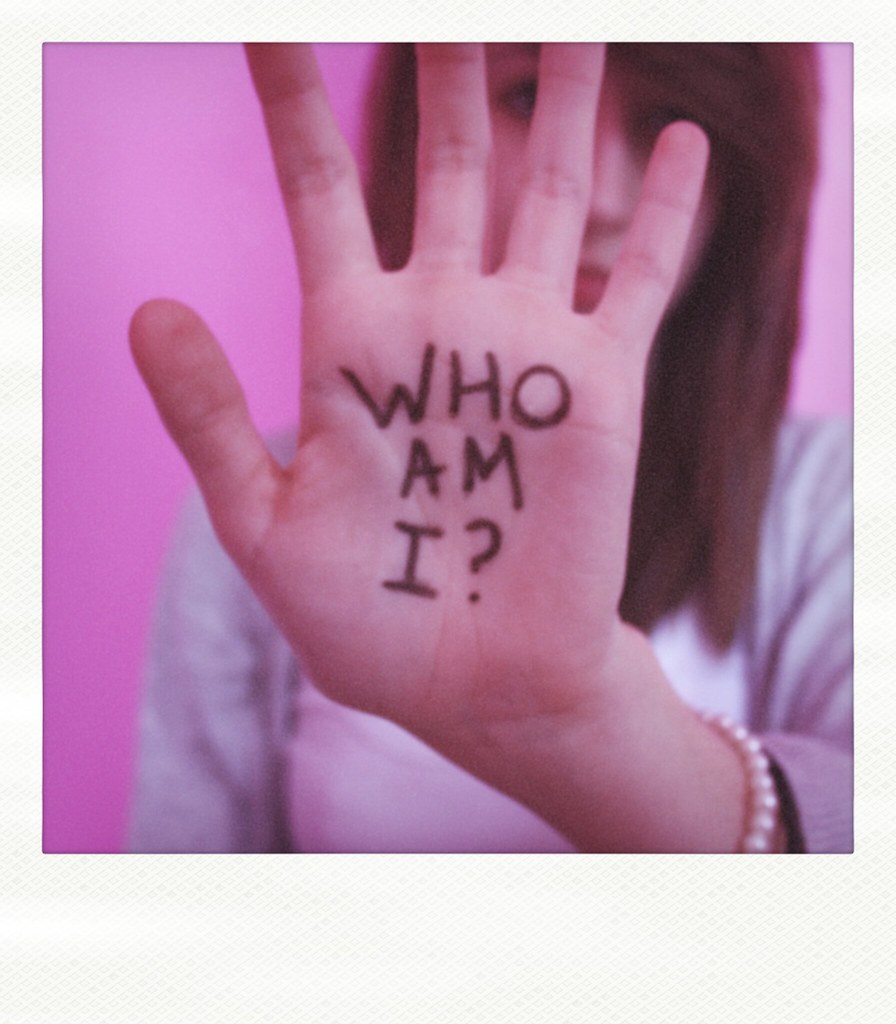 A woman dressed in a grey cardigan over a pale pink top against a darker pink background raises her left hand in front of her face. Across the palm is written  in Capitals in black, "WHO AM I?" 
