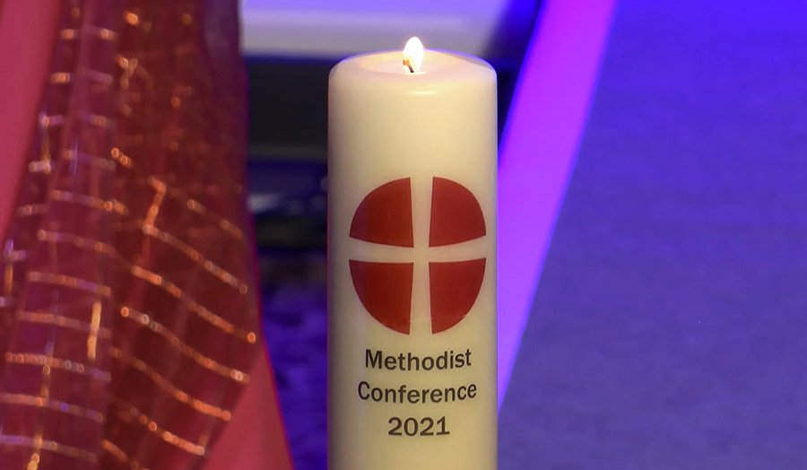 An off-white candle with the symbol of the Methodist Church, a withe cross on a red circle, and the wording "Methodist Conference 2021."