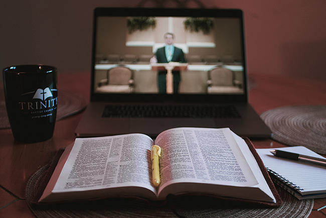 An open bible and a notebook and pen are in the foreground in front of a laptop on which is a picture of a church sanctuary in which a man in protestant robes appears to be preaching.robes is seen 