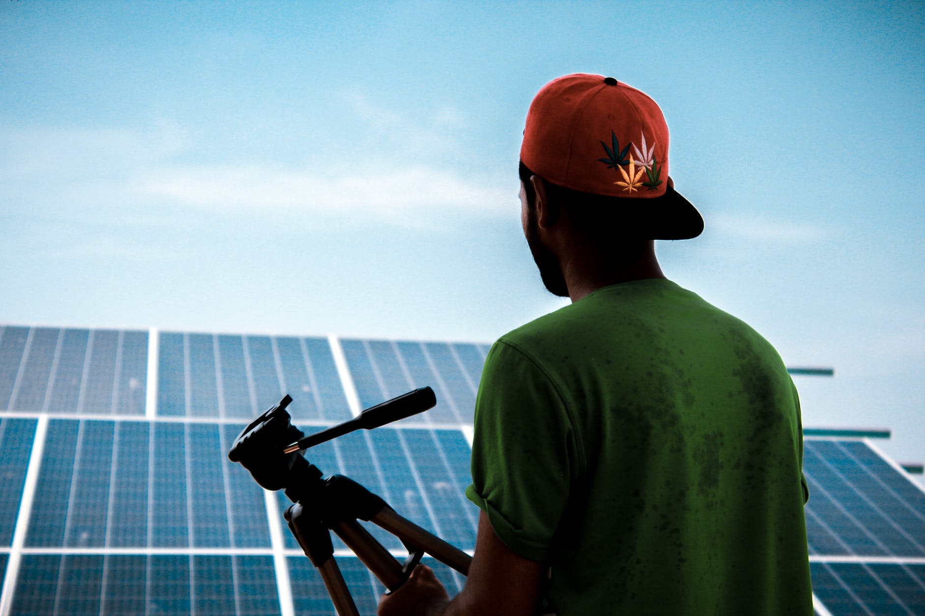 A man in a backwards baseball cap holding a tripod stands in front of an array of solar panels.