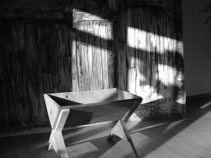 A wooden manger stands on a wooden floor. The light from the window casts a shadow of a cross on the wall behind.