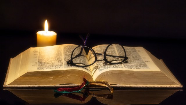 A pair of reading glasses lie on an open Bible illuminated by a single candle.
