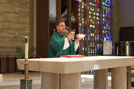 A male priest in dark green vestments blesses the communion cup at an altar with a stained glass window in the background.