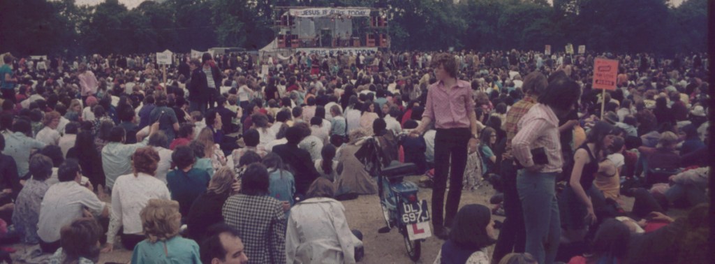 A small crowd of people sit on the grass in a park facing a stage.
