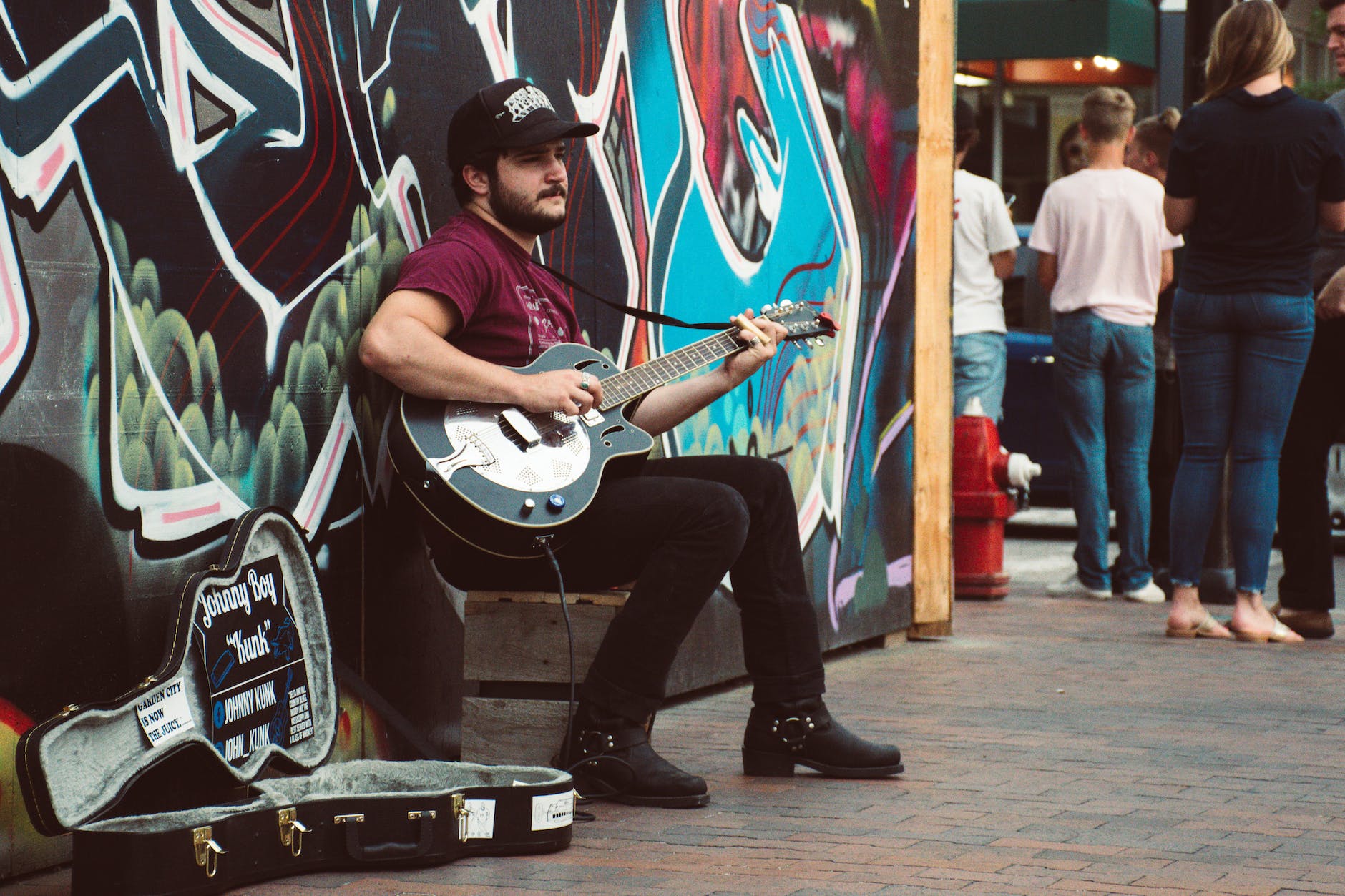 A busker playing a resonator guitar with a slide sitson the pavement in fromt of a graffitied wall.