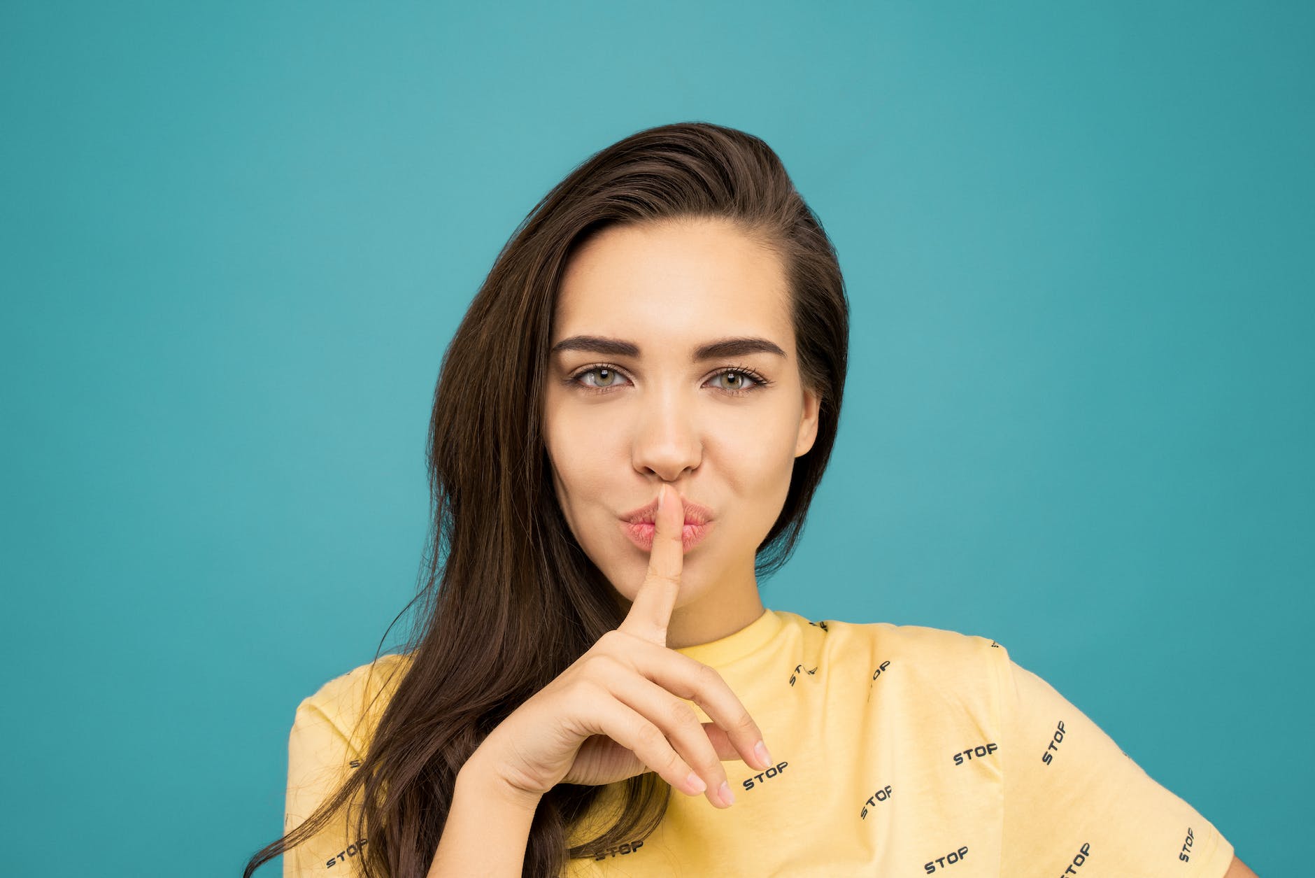 A woman in a yellow top containing the word 'stop' holds her finger to her mouth in a gesture of silence.