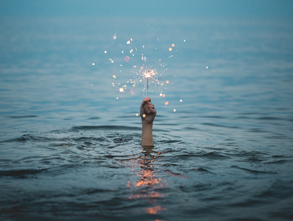 Our of a calm ocean a single hand is raised holding a sparkler firework.