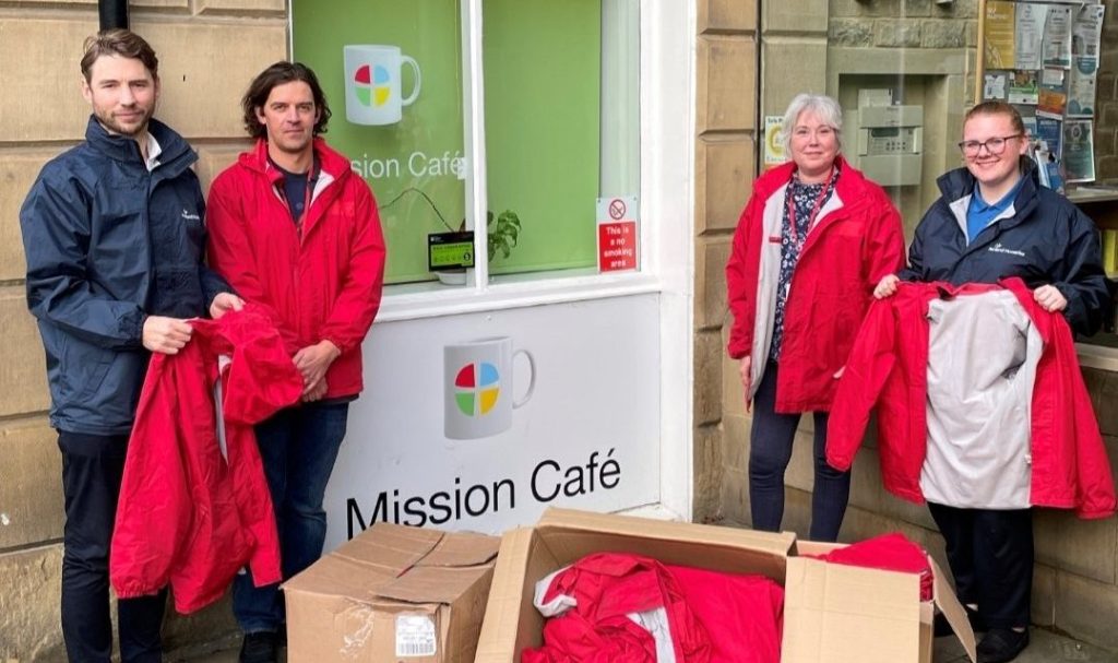 Four peole, two wearing and two holding red coats, stand by two boxes of coats outside the Mission Cafe, Huddersfield.