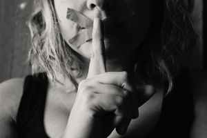 A woman with her mouth taped up holds a finger to her mouth to say shh.